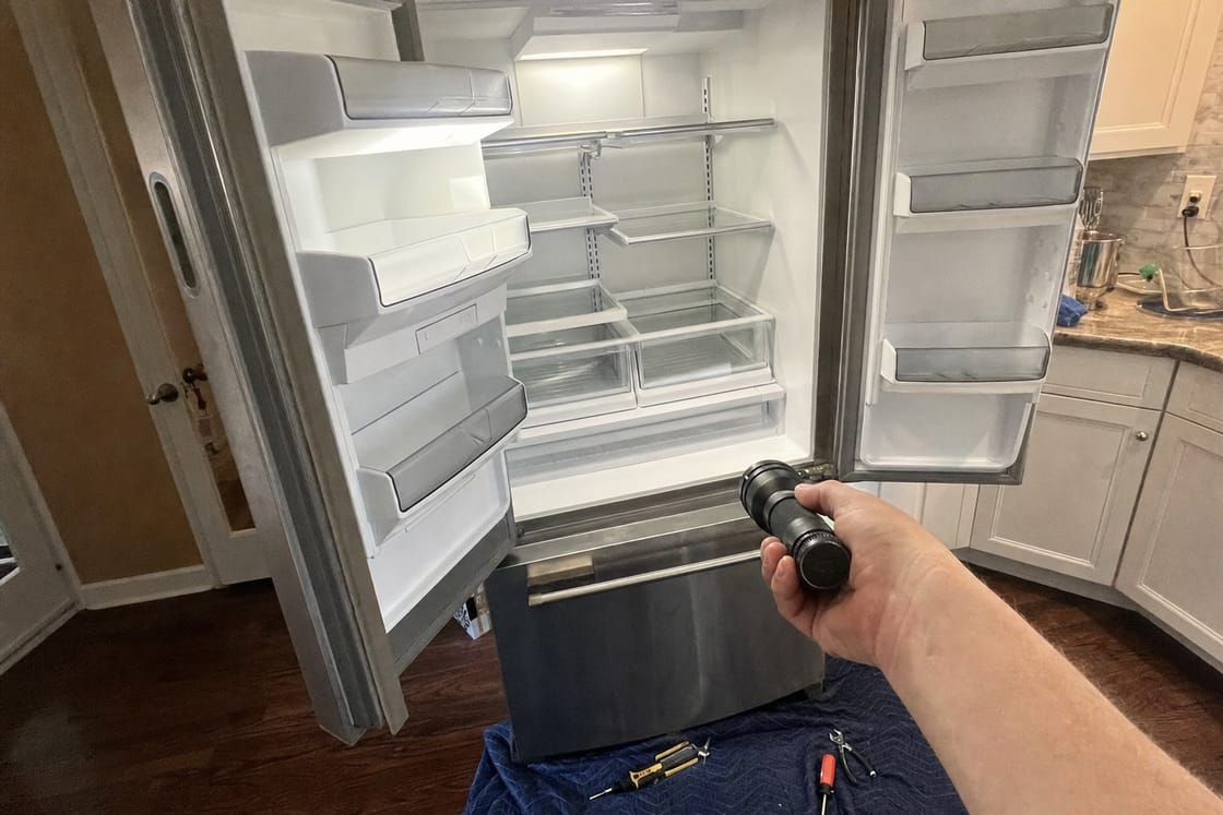 Built-in refrigerator standing open in a kitchen with the interior lit and a flashlight held on the empty shelves