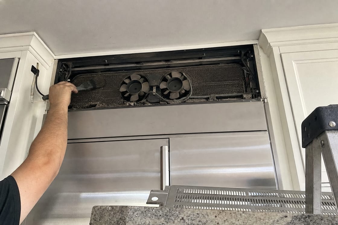 Upper grille panel of a built-in refrigerator held open, showing the condenser coil and its two fans