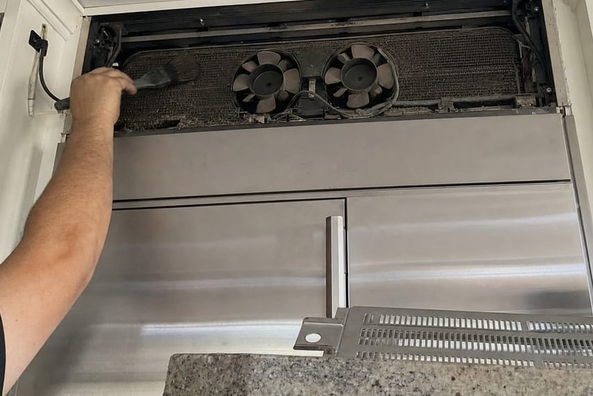 Hand holding open the upper grille of a built-in refrigerator with the condenser fans and coil behind it