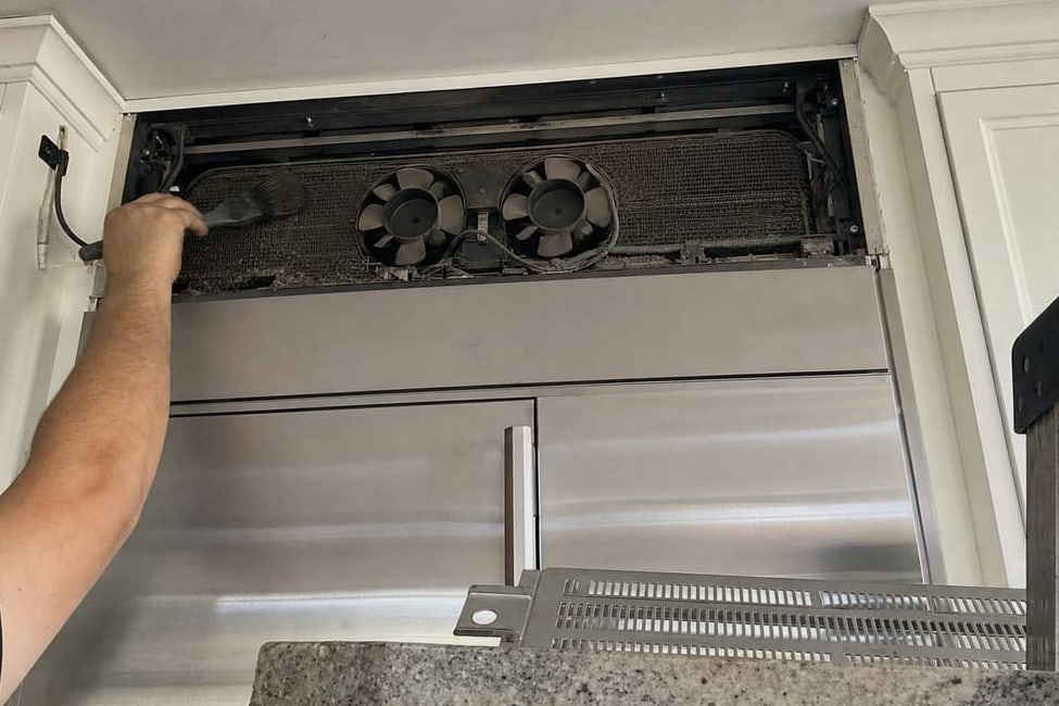 Condenser coil and twin fans behind the upper grille of a built-in refrigerator, the panel held open by hand