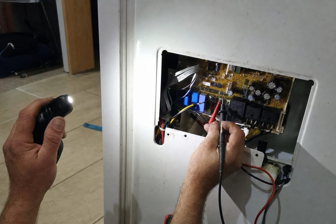 Control board exposed in its housing with a multimeter probe held on one of its terminals