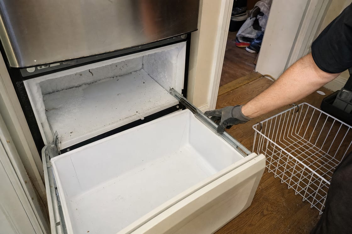 Freezer drawer pulled fully open on a built-in unit, wire basket alongside and a gloved hand on the drawer front