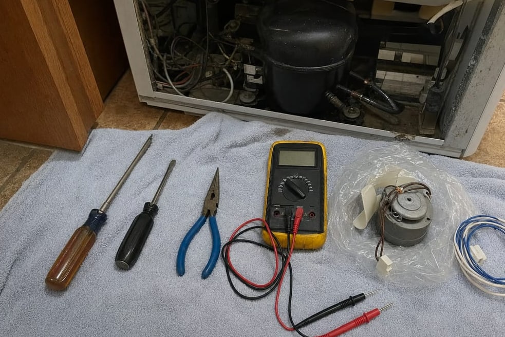 Multimeter and hand tools on a mat in front of a built-in refrigerator with the compressor compartment open
