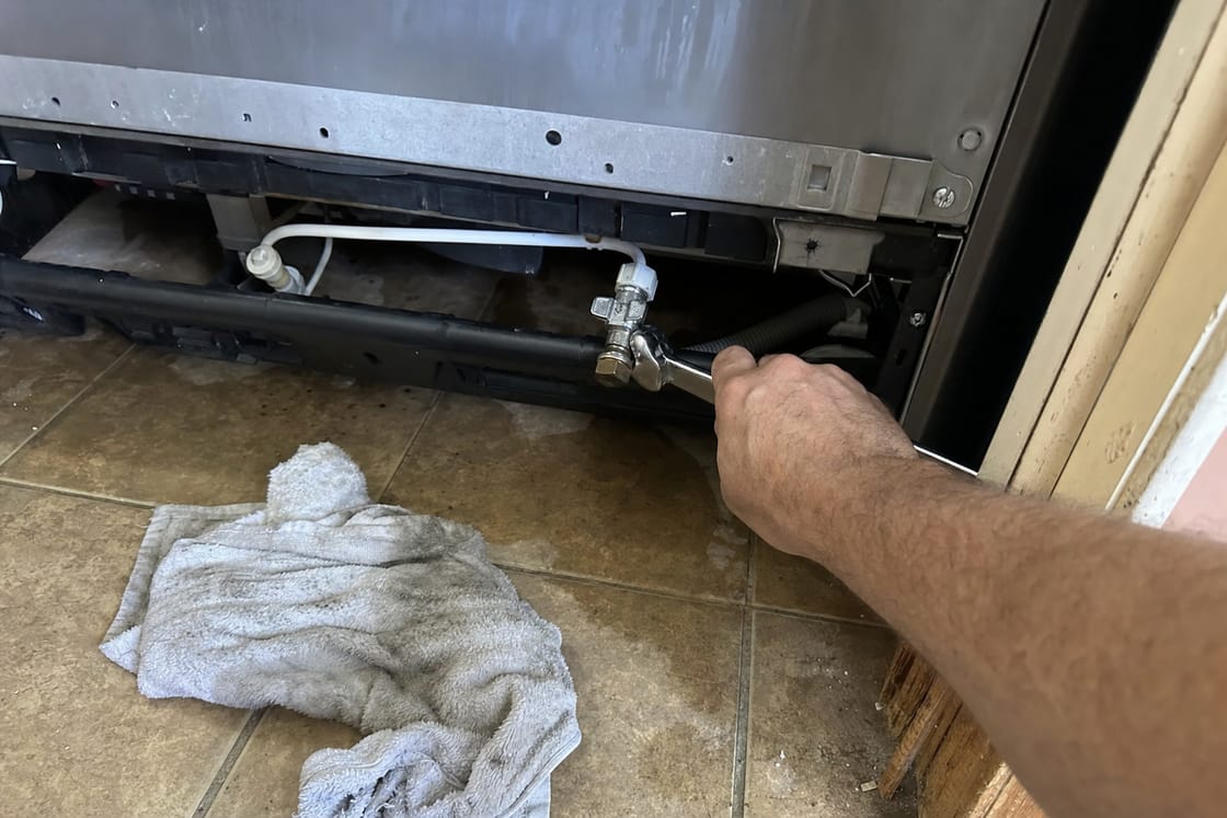 Wrench on the water shut-off valve and supply line at the base of a built-in refrigerator, a towel on the tile floor